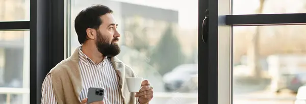 A handsome elderly man in casual attire holds a coffee cup and looks outside with reflection. — Stock Photo