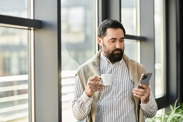 Relaxed senior man savors a warm cup of coffee while checking his smartphone by a large window — Stock Photo