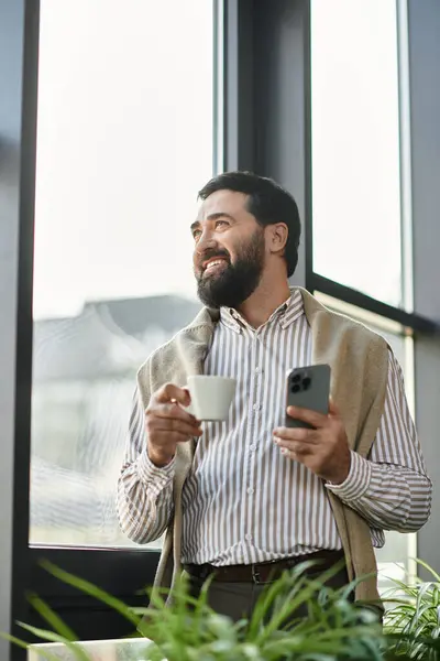 Senior gentleman stands by the window, savoring a cup of coffee and engrossed in his phone. — Stock Photo