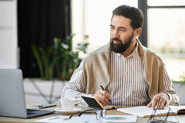 Casually dressed man with a beard sits at a desk, jotting down notes while focused on a laptop. — Stock Photo