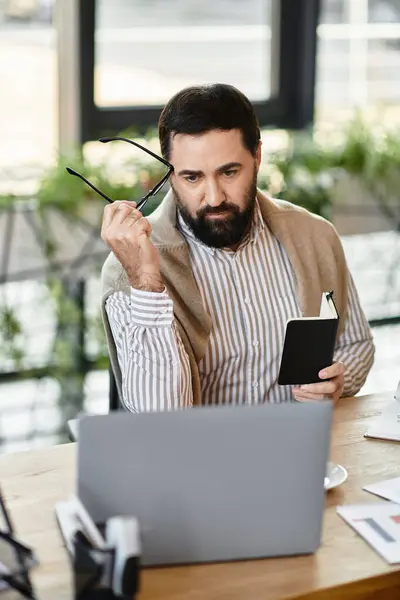 Dapper senior man with glasses uses a laptop and holds a notebook in a modern office. — Stock Photo