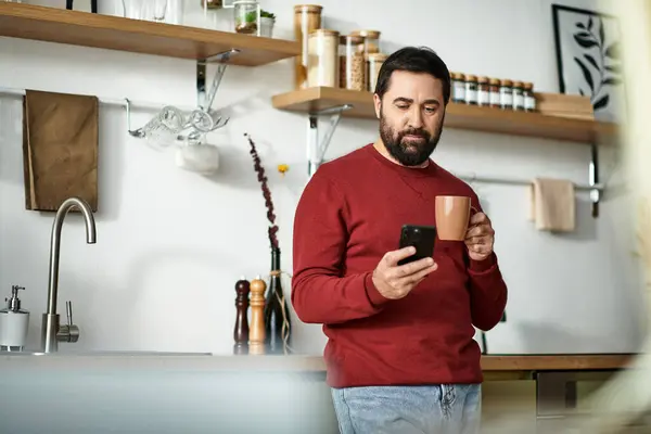 A handsome senior relaxes in a modern kitchen, sipping a warm drink and looking at his phone. — Stock Photo