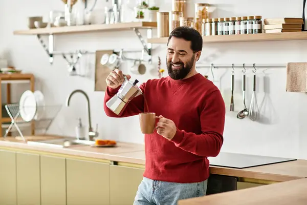 Un apuesto anciano felizmente vierte café caliente en una taza mientras sonríe en una cocina moderna. - foto de stock