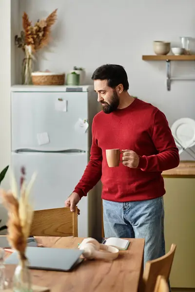 El hombre saborea su café de la mañana, de pie en una cocina cálida y acogedora adornada con una decoración sencilla. - foto de stock