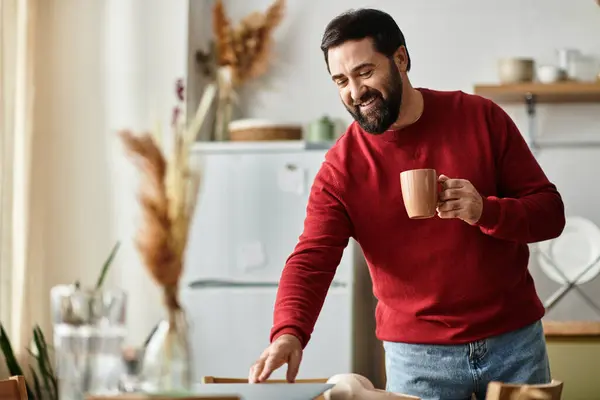 Hombre mayor guapo con atuendo casual sonríe mientras pone la mesa para una comida en una cocina caliente. - foto de stock