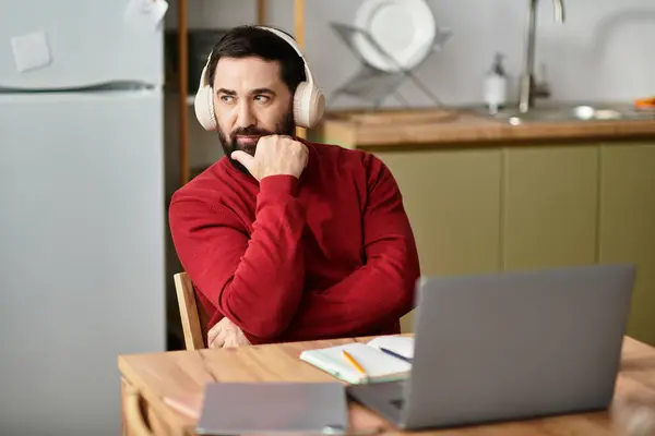 An elderly man listens to music with headphones while reflecting at a table in a bright kitchen. — Stock Photo
