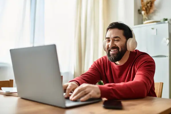 Cheerful older man listens to music with headphones while typing on his laptop at home — Stock Photo