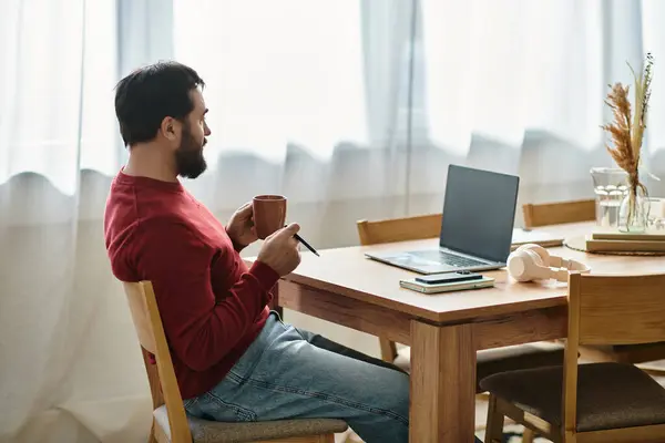 A handsome elderly man in casual wear sits at a wooden table, enjoying a warm drink while working. — Stock Photo