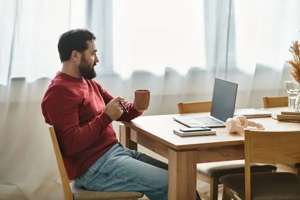 Casually dressed senior man with a warm drink focuses on his laptop at a sunny home workspace. — Stock Photo
