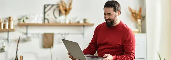 Ein attraktiver älterer Herr in lässiger roter Kleidung lächelt, während er seinen Laptop erkundet und dabei Wärme ausstrahlt. — Stockfoto