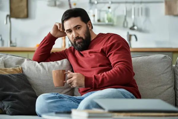 Handsome senior man sits comfortably on a couch with a warm beverage, reflecting thoughtfully. — Stock Photo