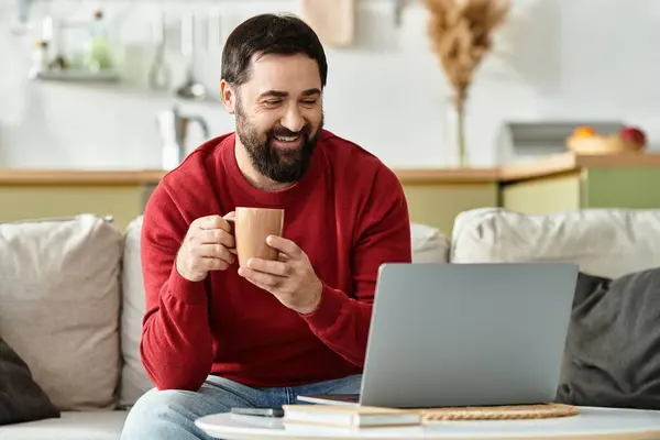 A gentle man relaxes on a couch, sipping coffee and engaging with his laptop. — Stock Photo