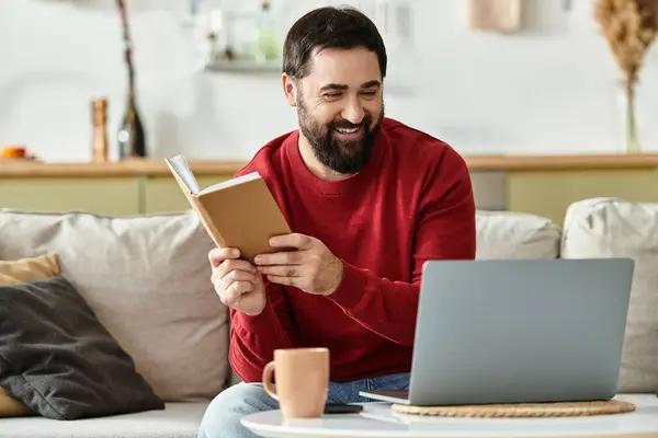 An elderly handsome man smiles as he reads a book, seated comfortably at home next to a laptop. — Stock Photo