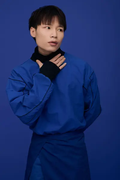 Stylish young Asian man stands confidently, hand on chest, amidst deep blue colors. — Stock Photo