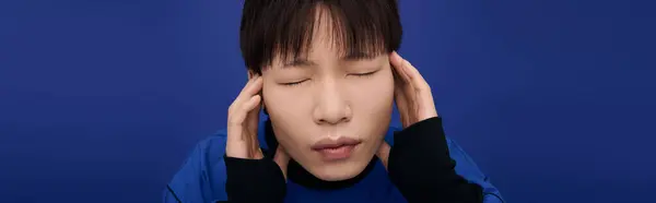 A handsome young Asian man with closed eyes looks deep in thought, set against a blue backdrop. — Stock Photo