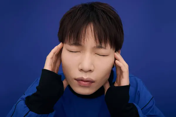 A young handsome asian man holds his head with closed eyes, conveying a sense of calmness. — Stock Photo