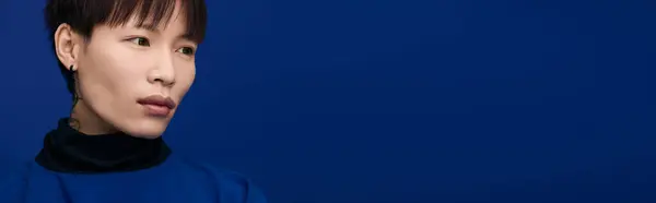 The young Asian man with unique style and good looks gazes thoughtfully against a blue backdrop. — Stock Photo