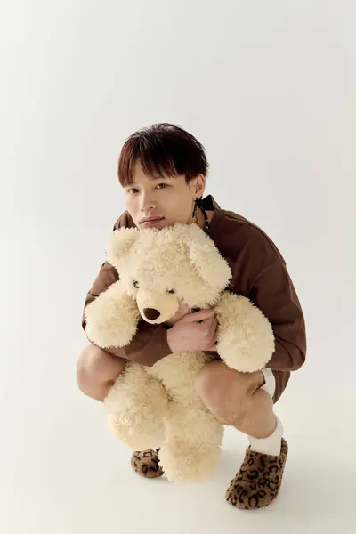 A young handsome asian man crouches down, softly embracing a fluffy teddy bear while smiling. — Stock Photo