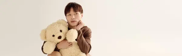Handsome young asian man smiles softly while holding a large teddy bear in a bright studio. — Stock Photo
