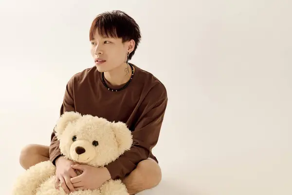 Un jeune et beau Asiatique est assis les jambes croisées avec un ours en peluche, profondément dans la pensée. — Photo de stock