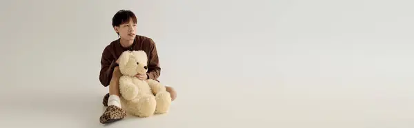 Dans un studio minimaliste, un beau jeune homme est assis avec un ours en peluche, rayonnant de chaleur et de confort. — Photo de stock