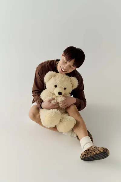 In a bright studio, a young handsome asian man smiles while hugging a large teddy bear warmly. — Stock Photo
