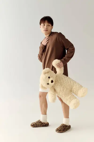 A young handsome asian man stands confidently, holding a teddy bear, dressed in casual loungewear. — Stock Photo