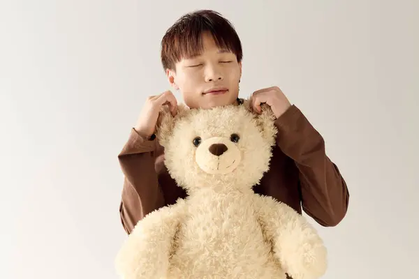 Vestido con un traje acogedor, un joven guapo asiático comparte un momento tierno con un lindo oso de peluche. - foto de stock