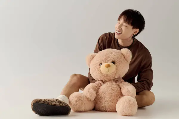 Un joven asiático sonríe en el suelo con un osito de peluche, añadiendo un ambiente juguetón. - foto de stock