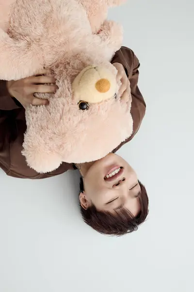 Un jeune homme asiatique joyeux se couche sur le dos, embrassant un ours en peluche géant, souriant joyeusement. — Photo de stock