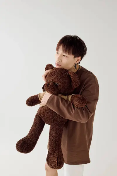 Handsome young asian man stands thoughtfully while hugging a soft teddy bear in a bright space. — Stock Photo