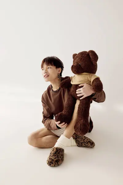 A handsome young Asian man sits on the floor, cuddling a teddy bear and daydreaming. — Stock Photo