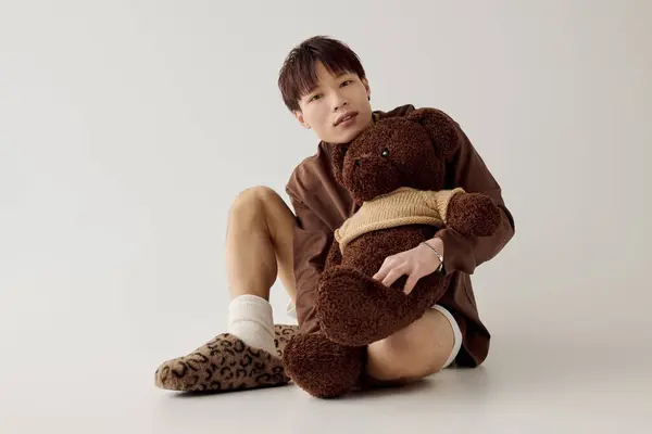 Un jeune homme asiatique charmant câline un ours en peluche avec un doux sourire tout en étant assis sur le sol. — Photo de stock