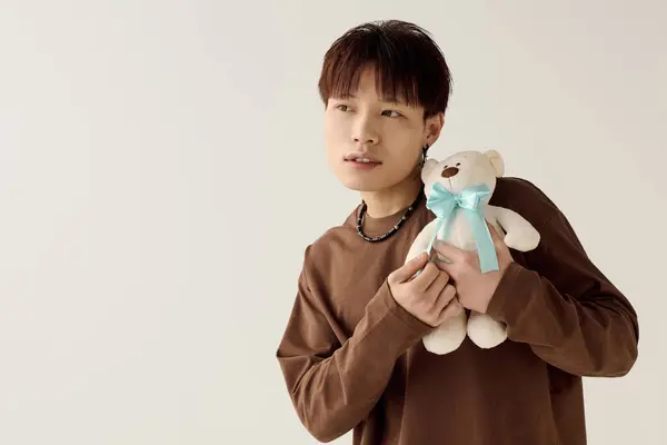 Un elegante joven asiático muestra su lado juguetón mientras sostiene un oso de peluche en un espacio brillante. - foto de stock