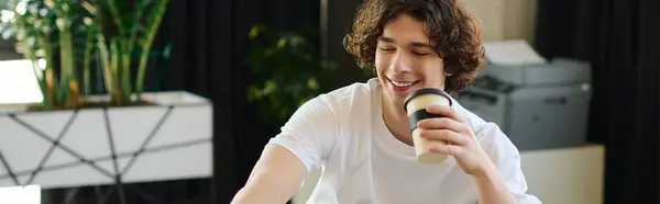 A young handsome man happily holds a coffee cup while focusing on work in a chic office setting. — Stock Photo