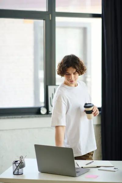Attractive young man stands in a bright office, glancing at his laptop while holding a coffee cup. — Stock Photo