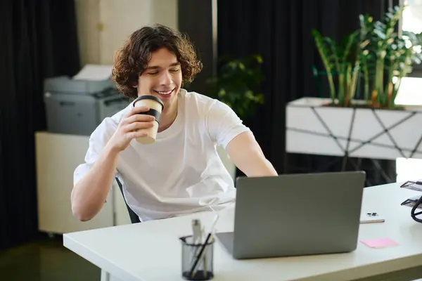 A handsome young man smiles as he sips his coffee at a sleek desk, focused on his laptop. — Stock Photo