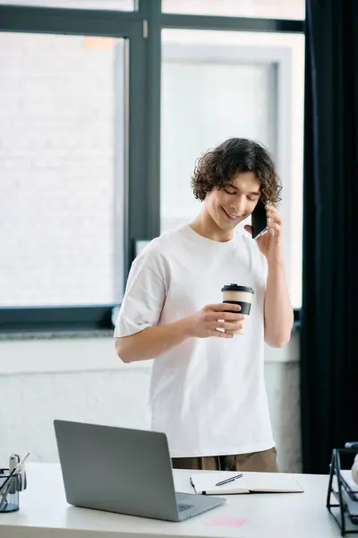 A young man stands in a cozy office, chatting on the phone while holding a coffee cup. — Stock Photo