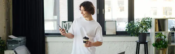A confident young man stands in a stylish office, coffee in hand, checking his phone. — Stock Photo