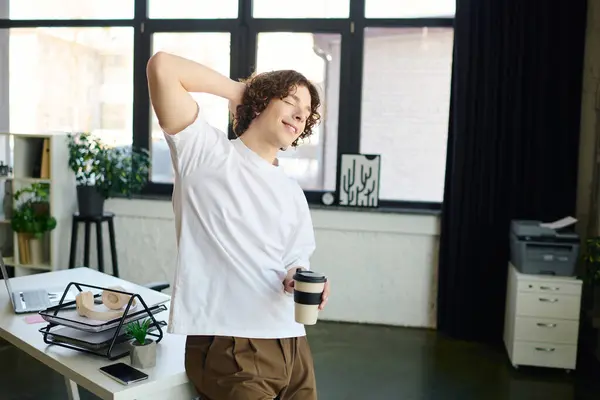 Guapo joven sonríe y se estira en su escritorio durante un refrescante descanso de café en la oficina. - foto de stock