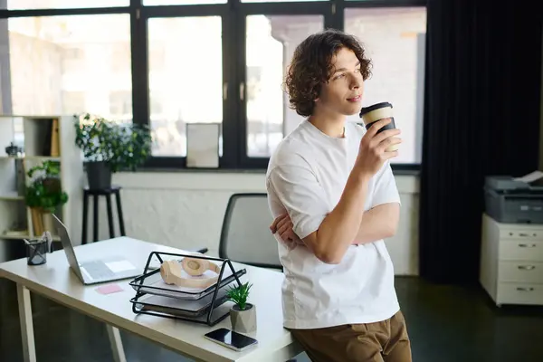 Attractive young man standing by his desk with a cup of coffee, lost in thought during work hours. — Stock Photo