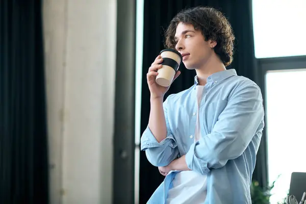 Young man in a blue shirt thoughtfully sips coffee while standing in a well lit office. — Stock Photo