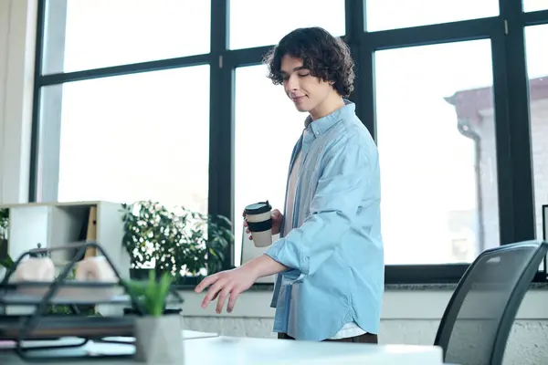 Young handsome man stands in a bright office holding coffee while reaching for a plant. — Stock Photo