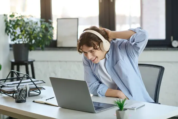 A young man in a stylish office happily interacts with his laptop while wearing headphones. — Stock Photo
