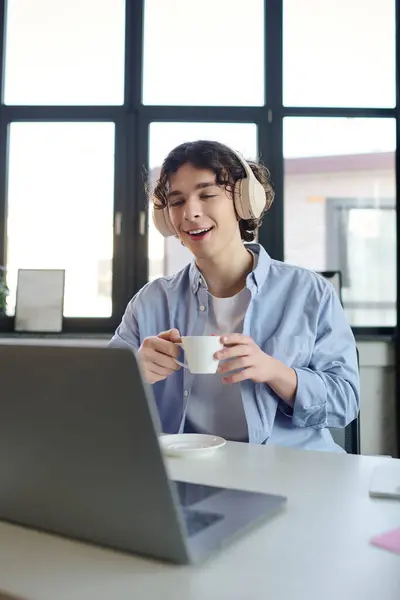 Sonriente joven sorbe café, centrado en su portátil en una oficina moderna llena de luz natural - foto de stock