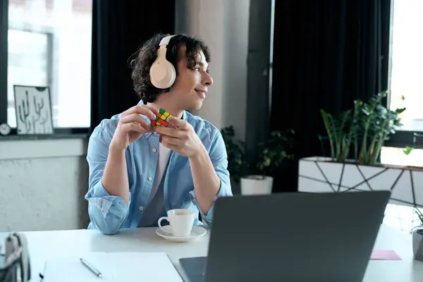 Um jovem bonito com fones de ouvido relaxa em seu escritório, resolvendo um Cubo Rubiks colorido. — Fotografia de Stock