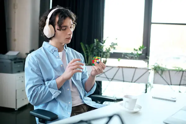 A focused young man wearing headphones works with a Rubiks cube in a stylish office setting. — Stock Photo