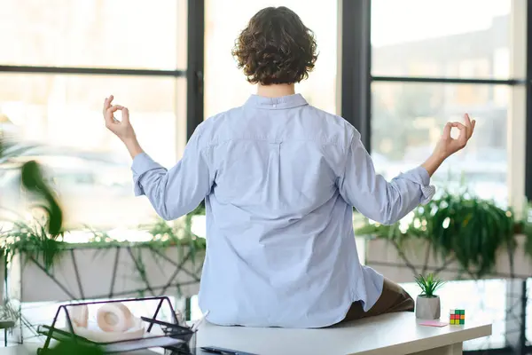 A stylish young man meditates cross legged on a desk, surrounded by greenery. — Stock Photo