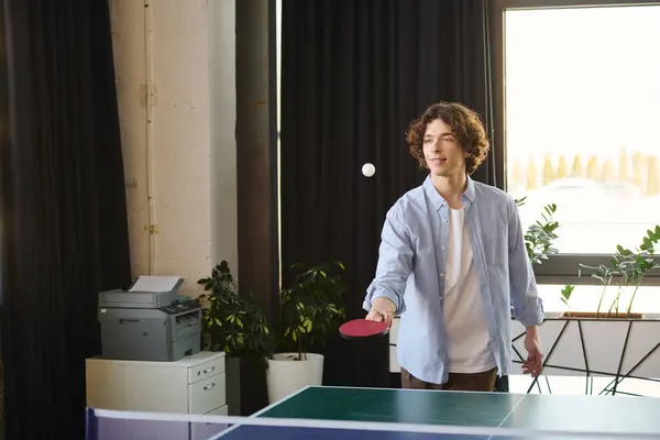 A young man with curly hair joyfully plays table tennis in a bright office. — Stock Photo