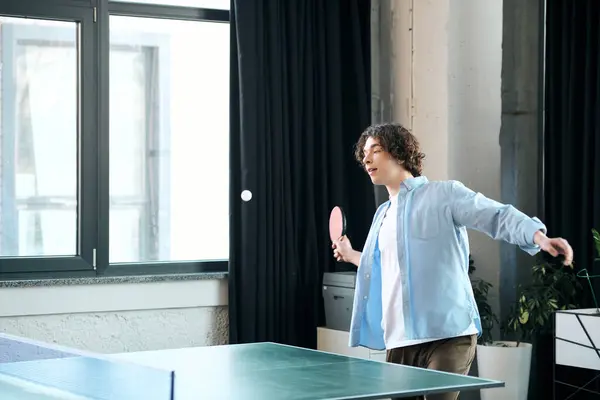 A young man with curly hair plays ping pong in a trendy office with large windows. — Stock Photo